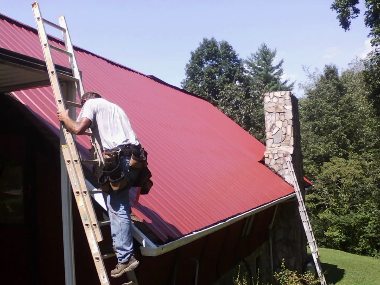 Roofer on a ladder beside a red metal roof and stone chimney — Soffit & Fascia Repair by Roofing Brothers in Weirton